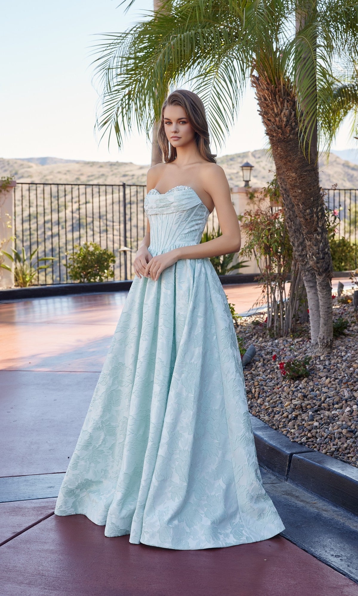 A woman stands outdoors by palm trees in the Long Prom Dress: Nox Anabel T1912, which features a jeweled sweetheart neckline, strapless bodice, and a full sage green skirt.